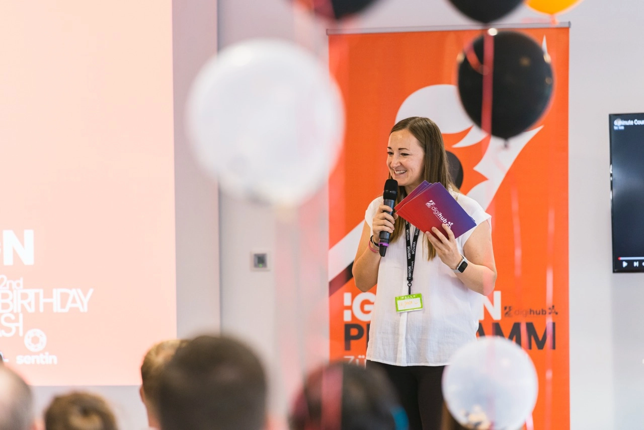 Woman holding a microphone and event booklets speaking in front of an audience with balloons in the foreground.