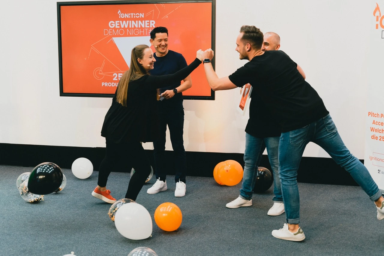 Two people fist bumping with balloons on the floor and a third person smiling in front of an orange screen reading 'Ignition Gewinner Demo Night'.