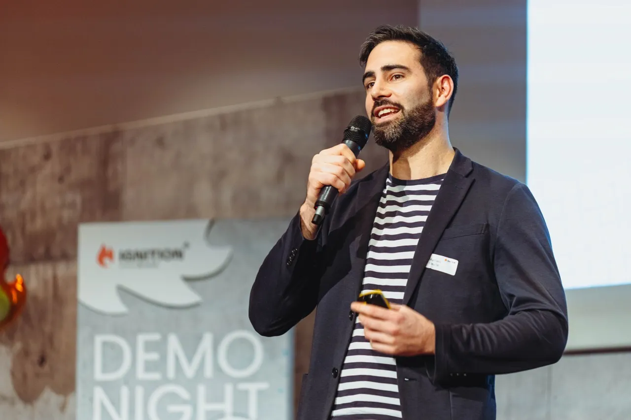 Man with beard speaking into a microphone at a demo night event.