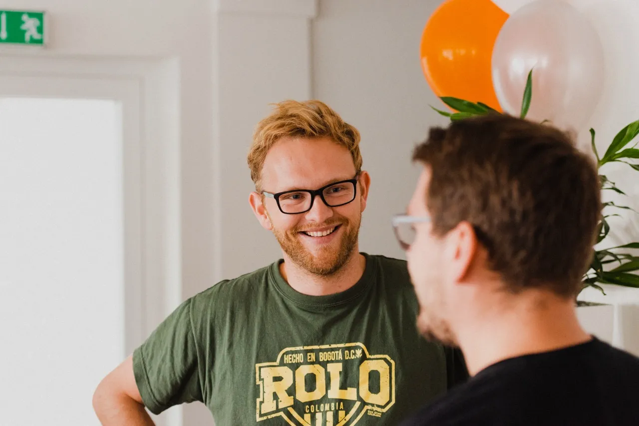 Smiling man with glasses and a green Rolo Colombia t-shirt talking to another man indoors with balloons in the background.