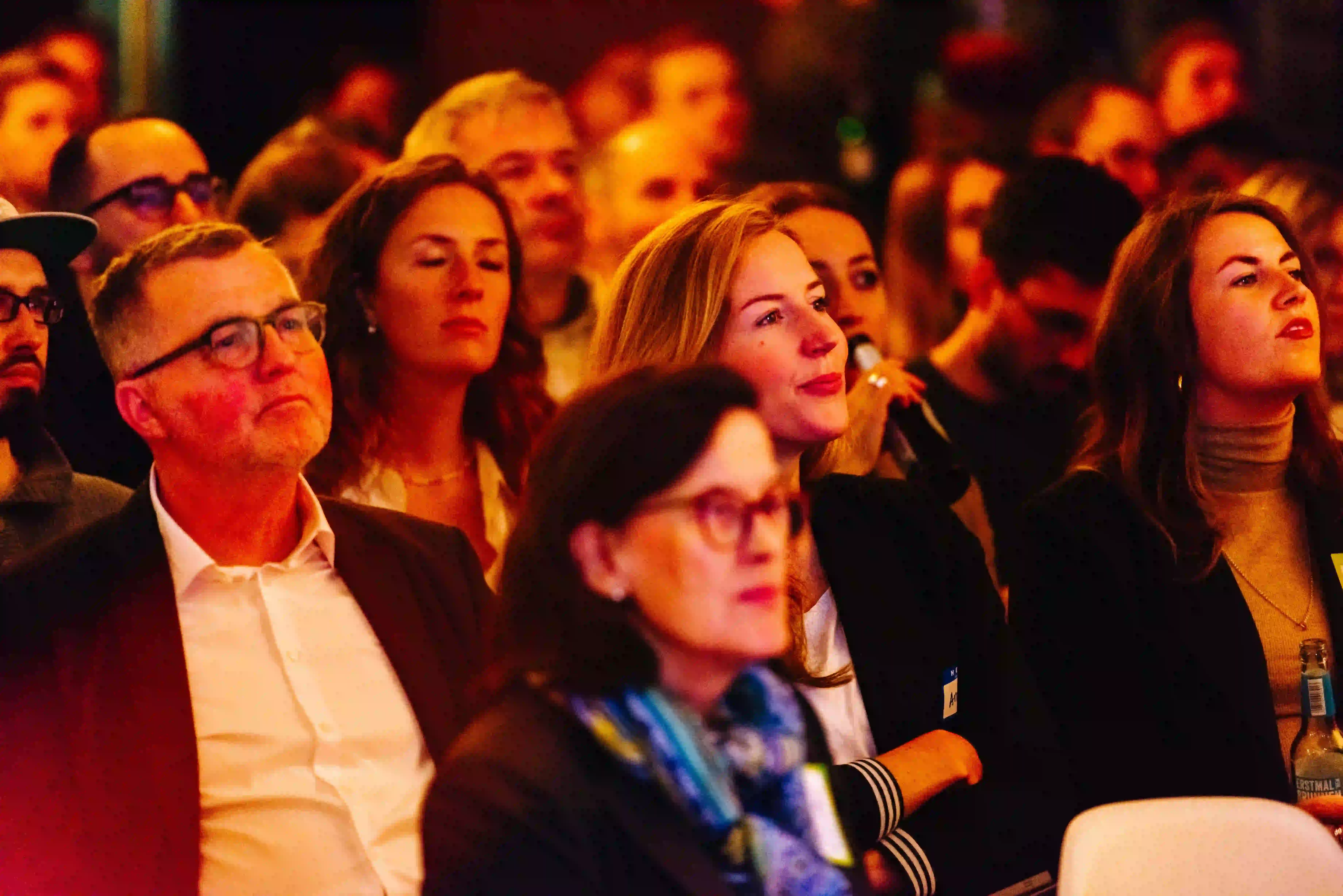 Audience of diverse adults attentively watching an event, some holding beverages in a dimly lit room.