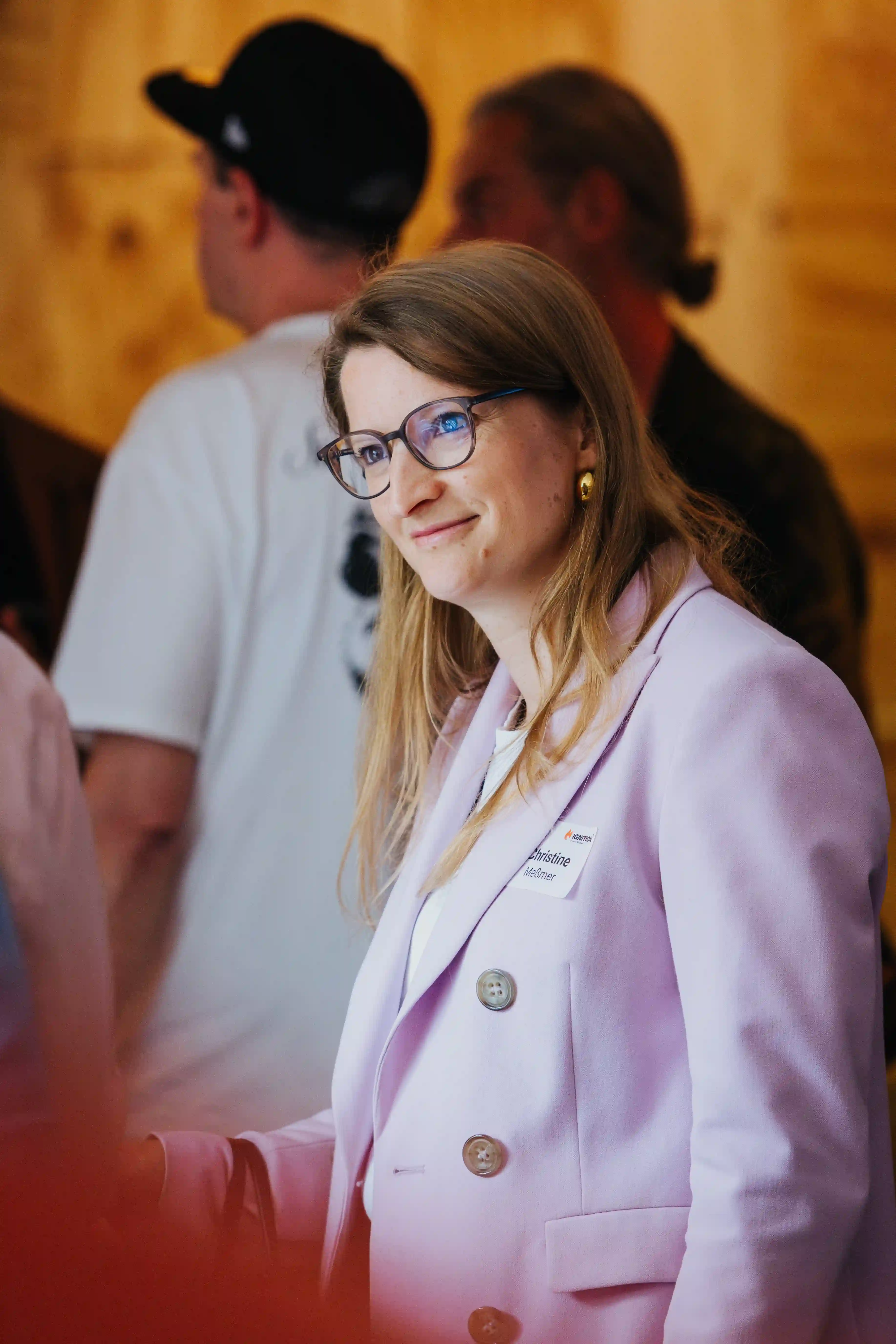 Woman with glasses and light brown hair wearing a lavender blazer with a name tag, smiling indoors.