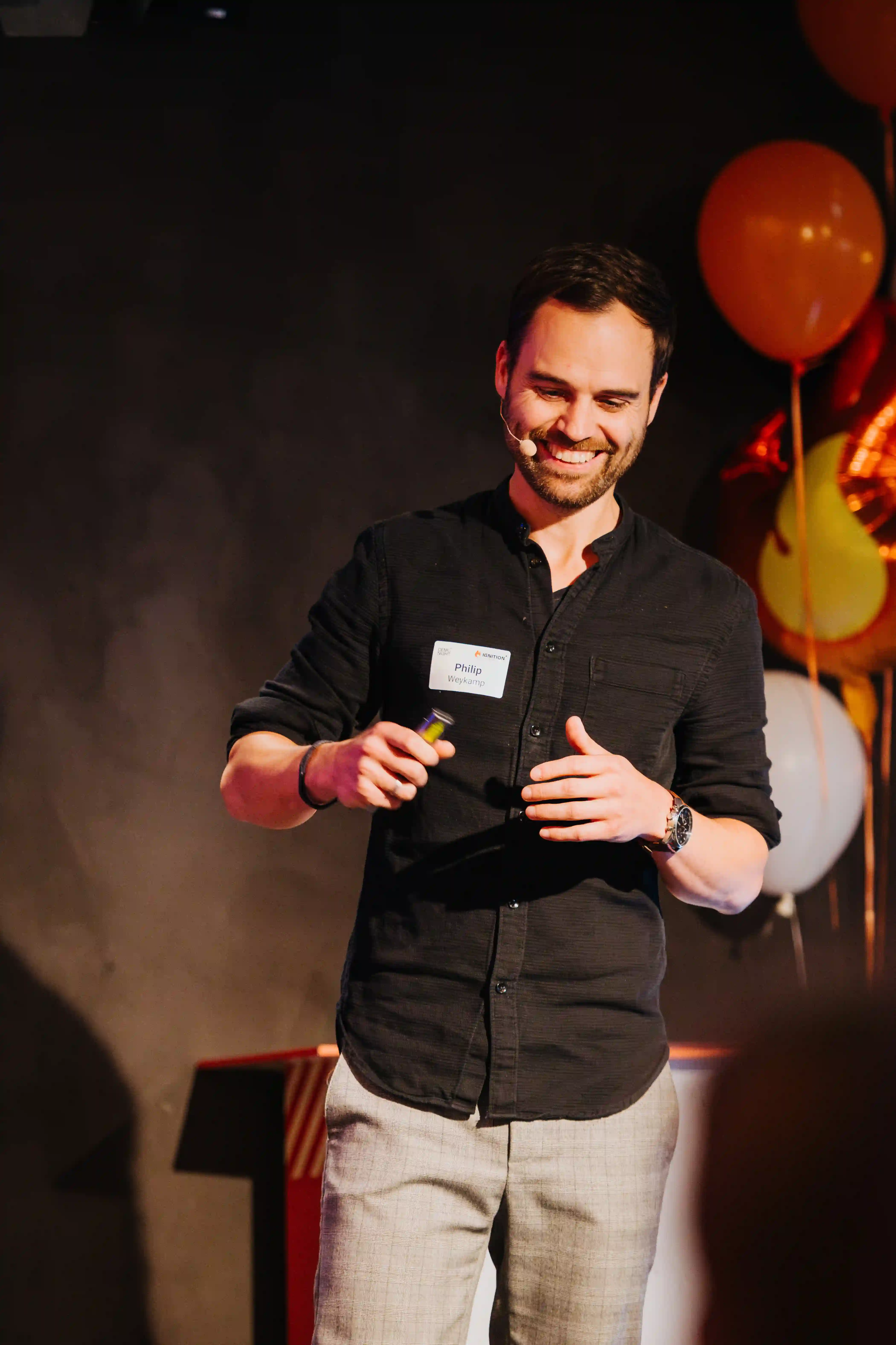 Smiling man named Philip Weykamp speaking with a headset microphone and holding a remote, standing in front of a dark background with balloons.