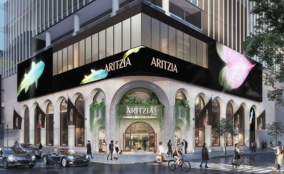 Architectural rendering of the planned Aritzia flagship boutique in the former Nordstrom space at CF Pacific Centre in downtown Vancouver, seen from a street corner. A curved stone facade with tall arched windows carries the Aritzia name over the entrance and on hanging banners, with a black digital display band above showing fish and flower imagery, and pedestrians, cyclists and cars on the street.