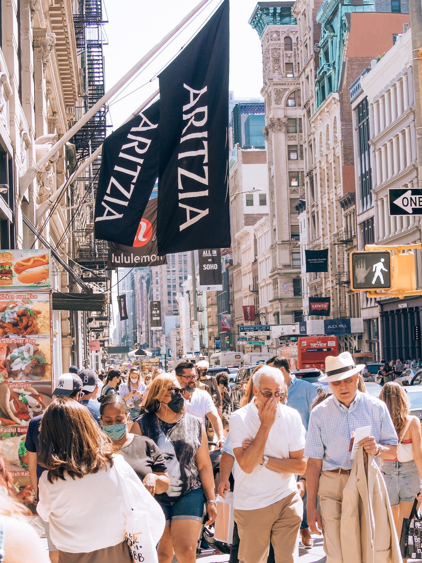 Street photograph of Broadway in SoHo, New York, on a sunny day. Two black flags reading ARITZIA hang over the sidewalk in the foreground, with a red and black Lululemon storefront banner just behind them, cast-iron buildings and shop signs including Levi's, Alex Mill and PacSun down the street, and a dense crowd of pedestrians below.