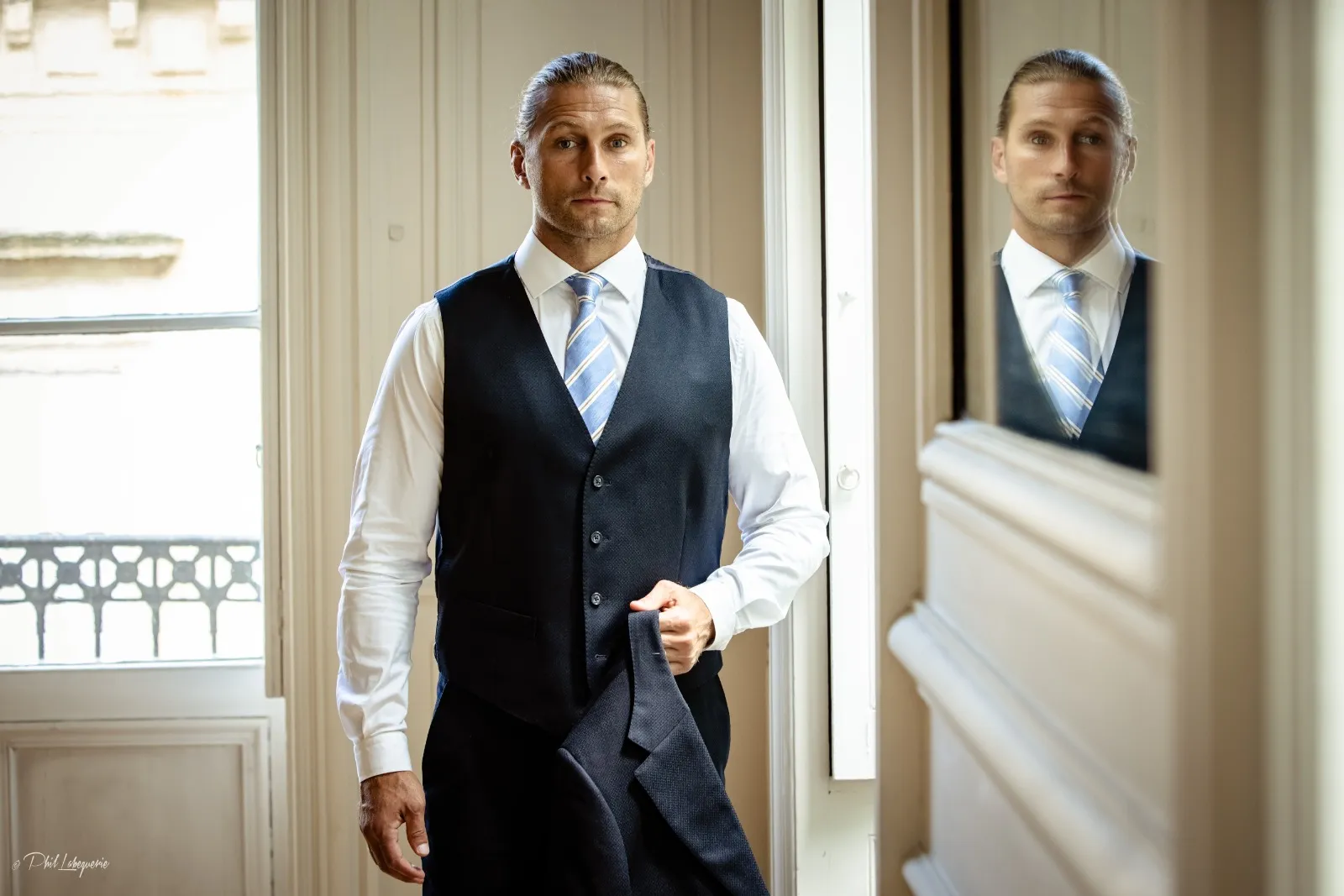 Homme portant un gilet de costume sur mesure bleu marine, chemise blanche et cravate rayée bleu clair, tenant sa veste à la main dans un intérieur lumineux — création Sartore Tailoring, Bordeaux.