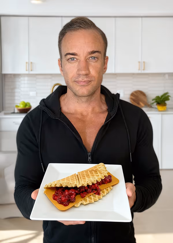 Man in black jacket holding a square white plate with two waffle halves filled with red berry compote in a bright kitchen.