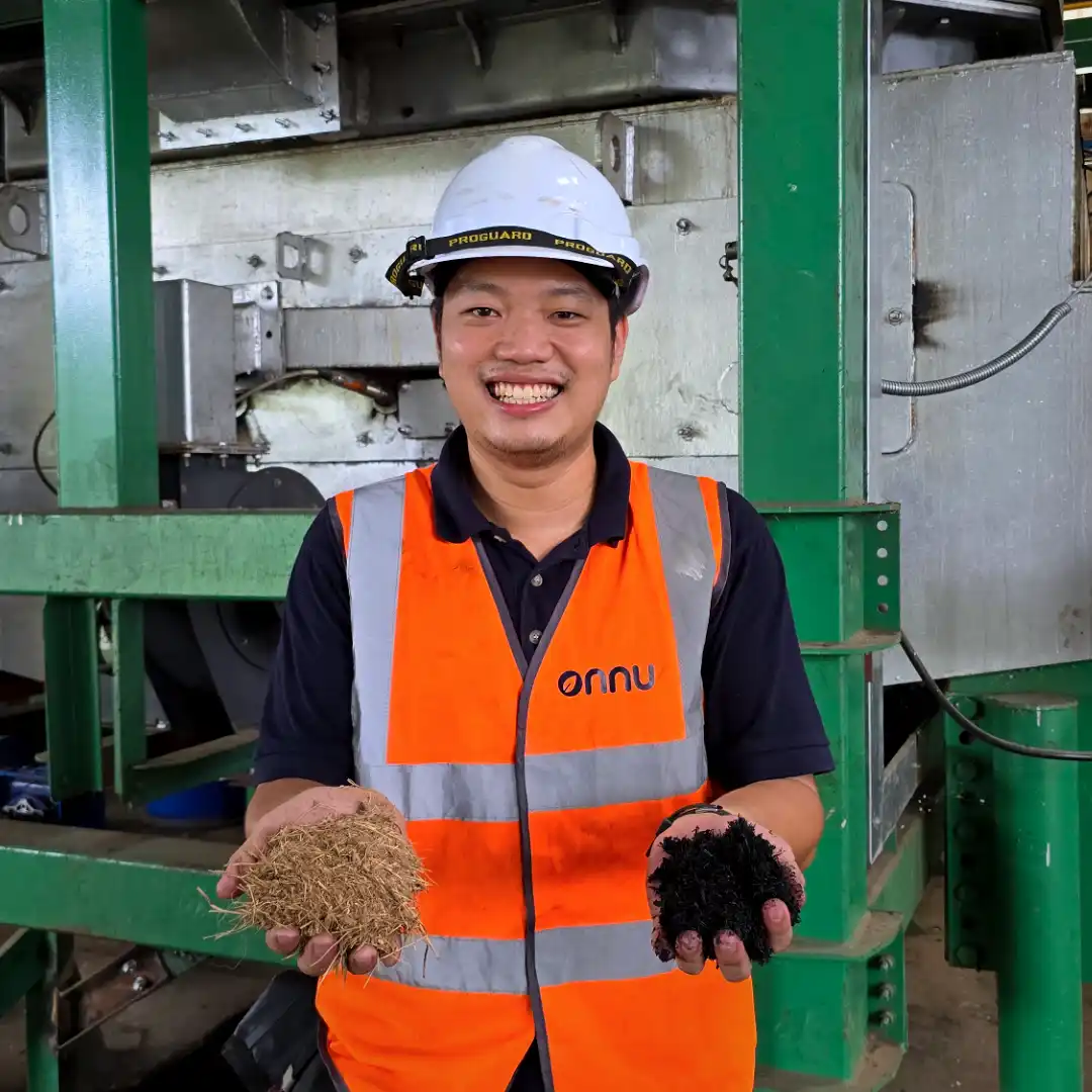 A person in hi-vis, in front of a pyrolysis machine, holding biochar in one hand and feedstock in the other