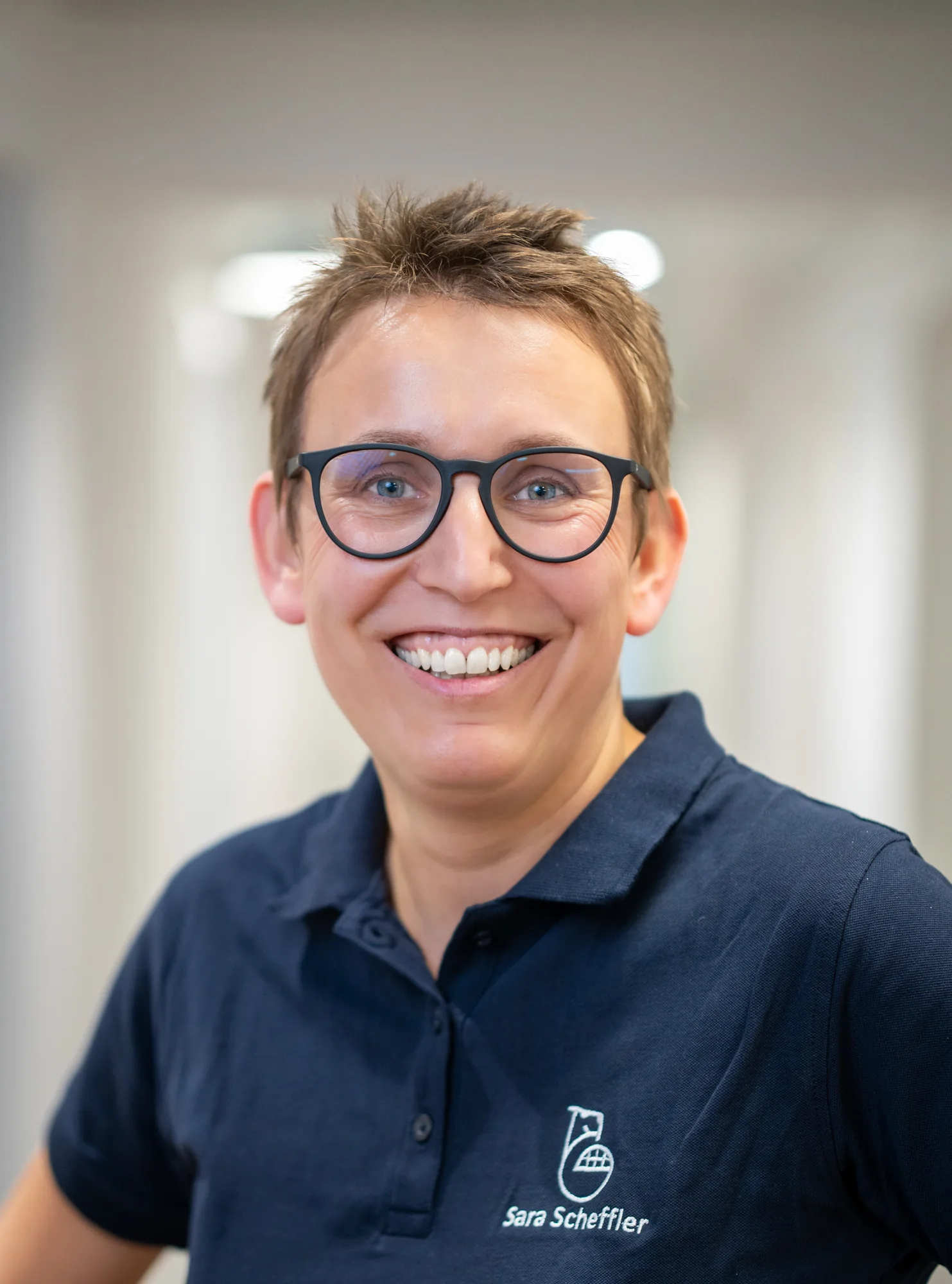 Frau mit kurzen braunen Haaren, Brille und blauem Polohemd mit Logo, lächelt in die Kamera.