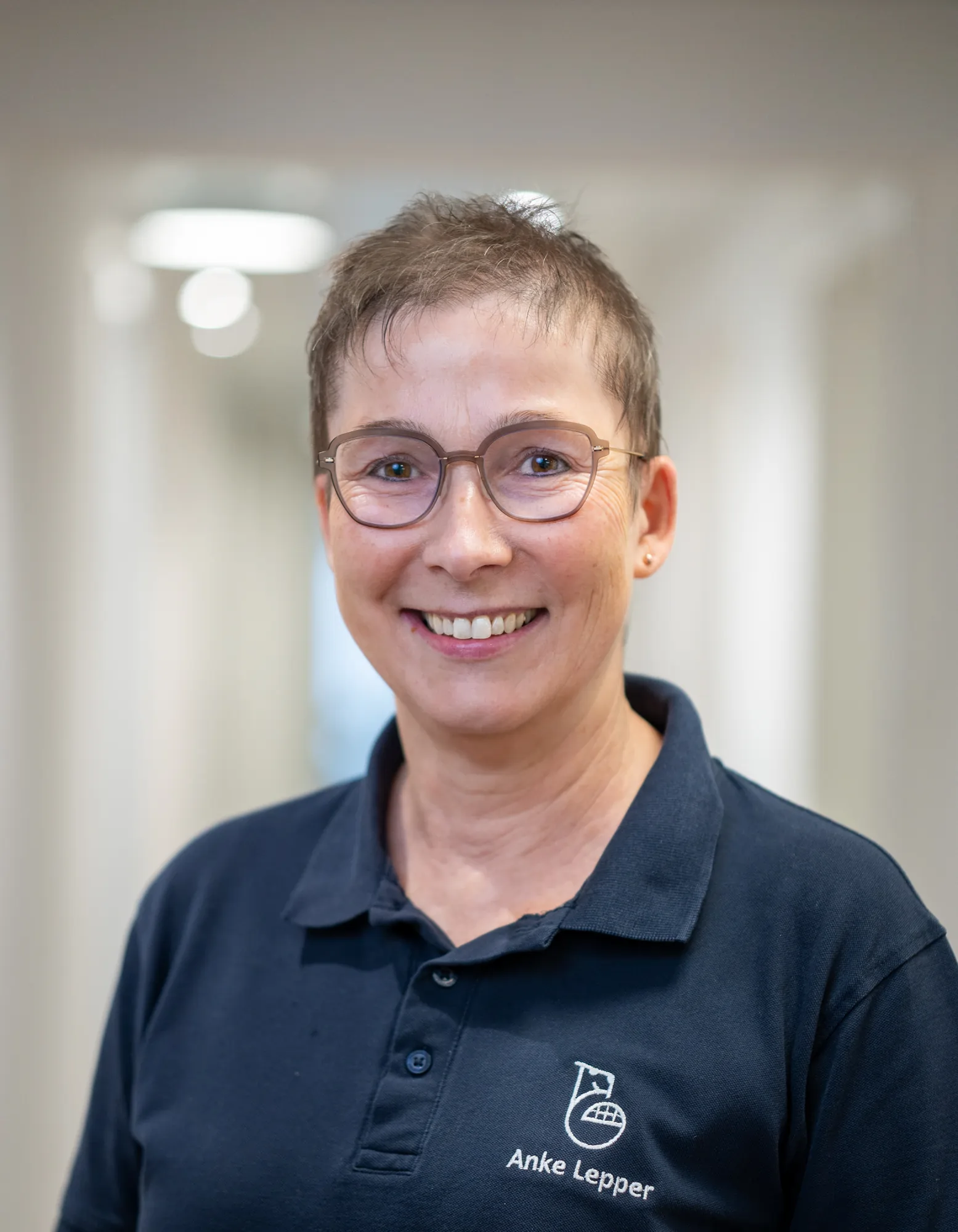 Porträt einer lächelnden Frau mit kurzen Haaren und Brille, die ein dunkelblaues Poloshirt mit der Aufschrift Anke Lepper trägt.