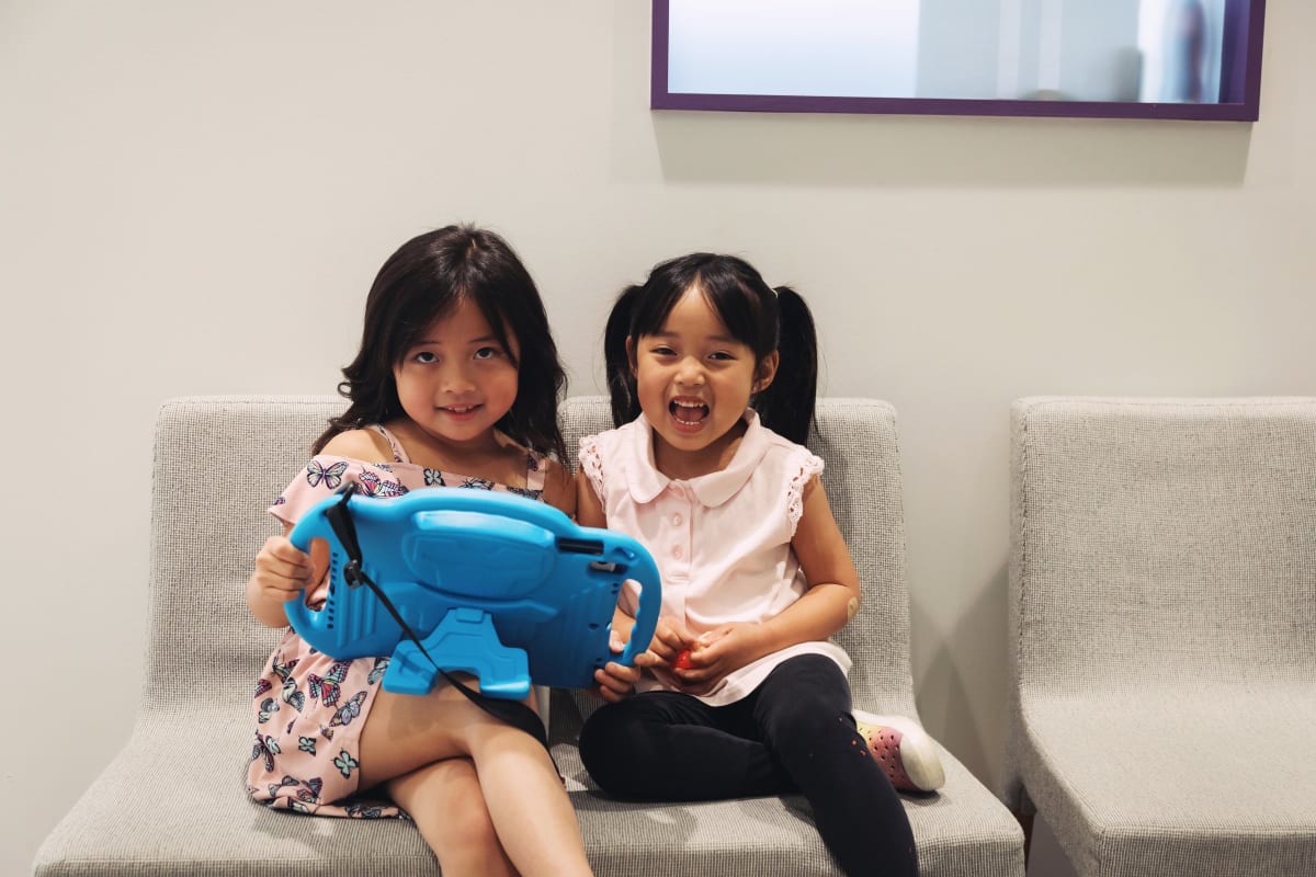 Two young children sitting together on a couch, one holding a blue book or tablet while reading