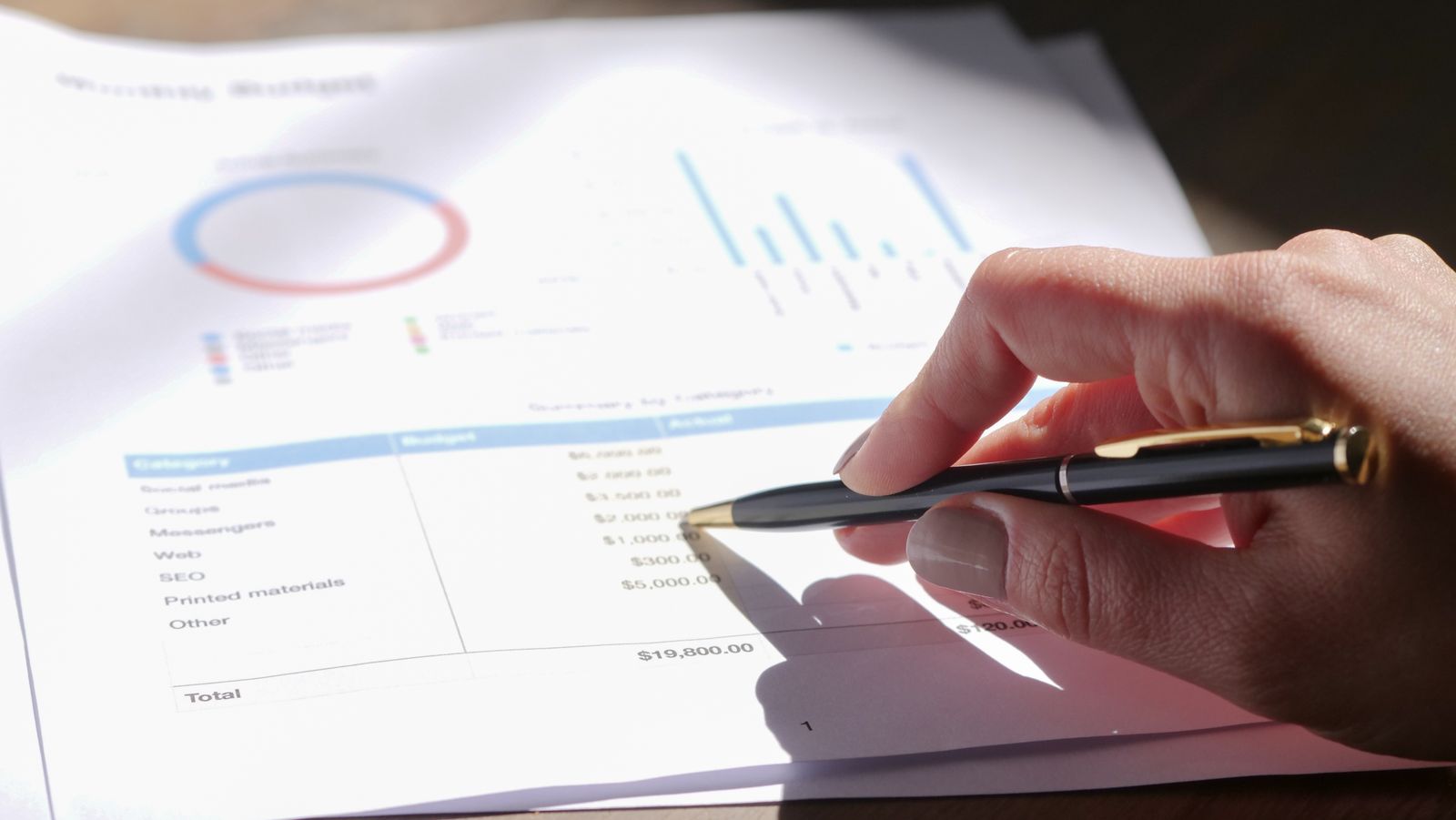 Hand with a pen reviewing a printed budget with charts