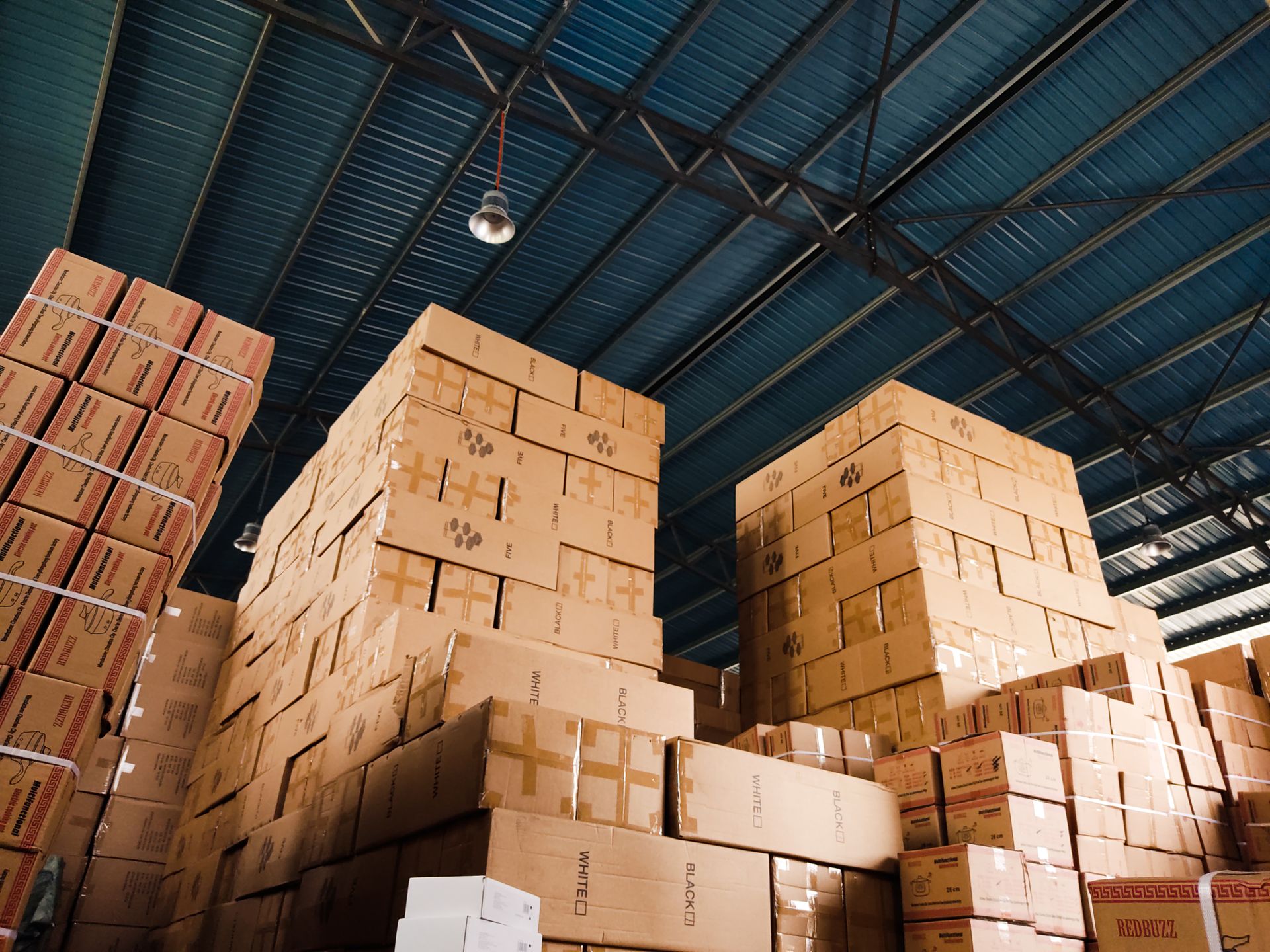 Tall stacks of cardboard shipping boxes inside a warehouse with a blue metal roof