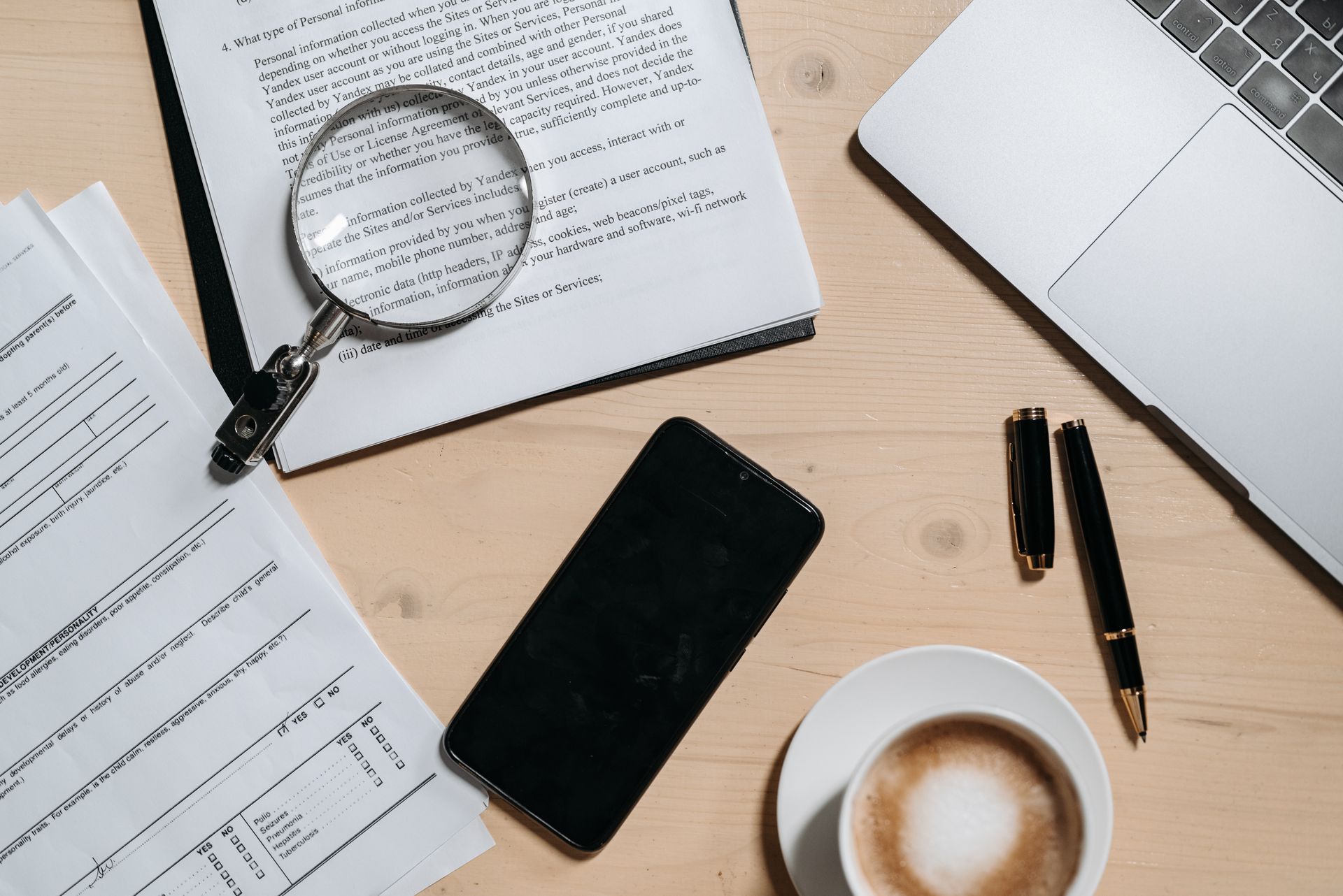 Printed documents under a magnifying glass beside a phone, pen and cup of coffee on a desk