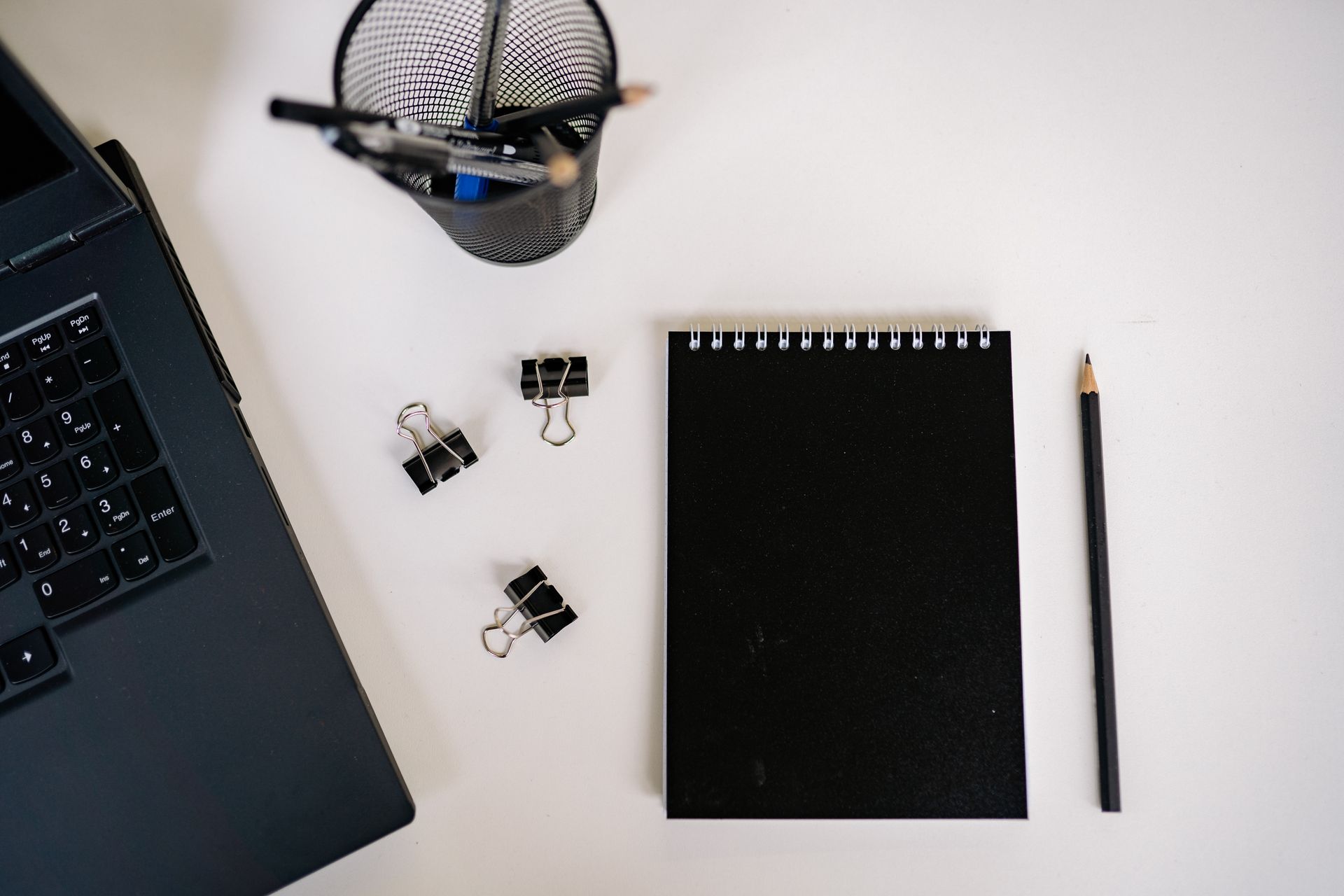 Overhead view of a laptop, black notepad, pencil and binder clips on a white desk