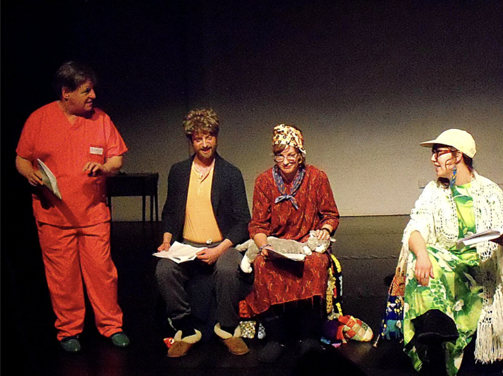 A scene from a 2013 performance of “Escape from Century Hills” by Steve Orth & Lindsey Boldt. From Left: Kevin Killian is standing, wearing pink scrubs. Steve Orth is seated, wearing a blue plaid bathrobe, gray sweats, slippers, and a wig. Lindsey Boldt is seated with a gray stuffed kitty in her lap, wearing a floor-length paisley mumu, flower print head scarf, and glasses. Sara Larsen is seated, wearing a yellow ball cap, green and yellow psychedelic print mumu, white crochet scarf, and glasses. All hold scripts.