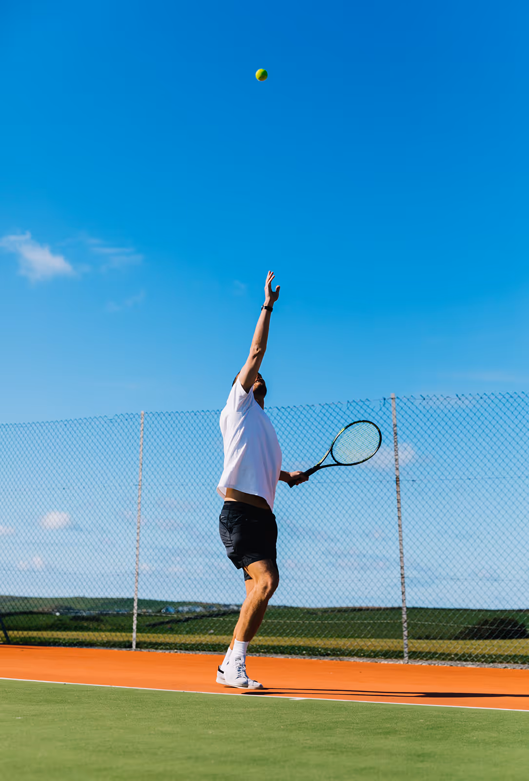 Male tennis player in white shirt and black shorts preparing to serve on outdoor court under clear blue sky.