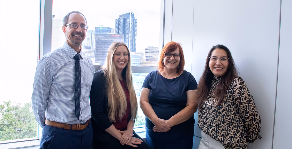 Four smiling professionals in business attire standing indoors with a cityscape through a window behind them.
