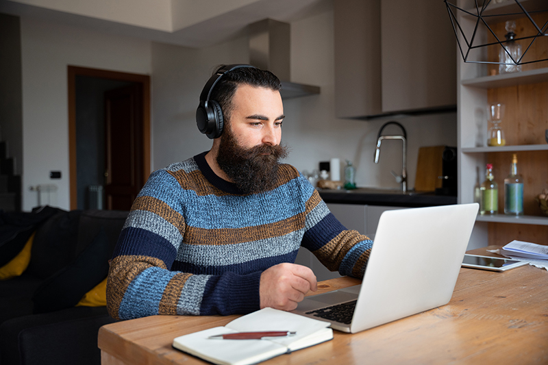 Bearded man with headphones sits at a kitchen table looking at an open laptop with a notebook resting open next to it