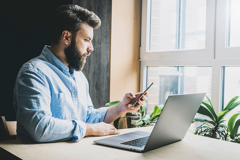 Bearded man at a desk by a window looking down at his phone while next to an open laptop