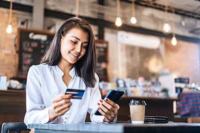 Woman holding a credit card and smartphone, sitting at a table in a café with a coffee cup nearby.