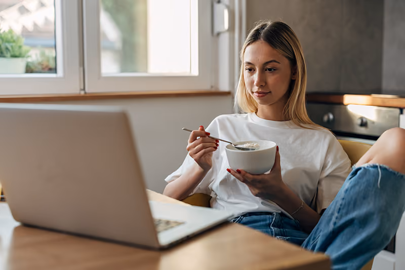 A young blonde woman holds a bowl of oatmeal with a spoon while seated in front of a laptop at a kitchen table
