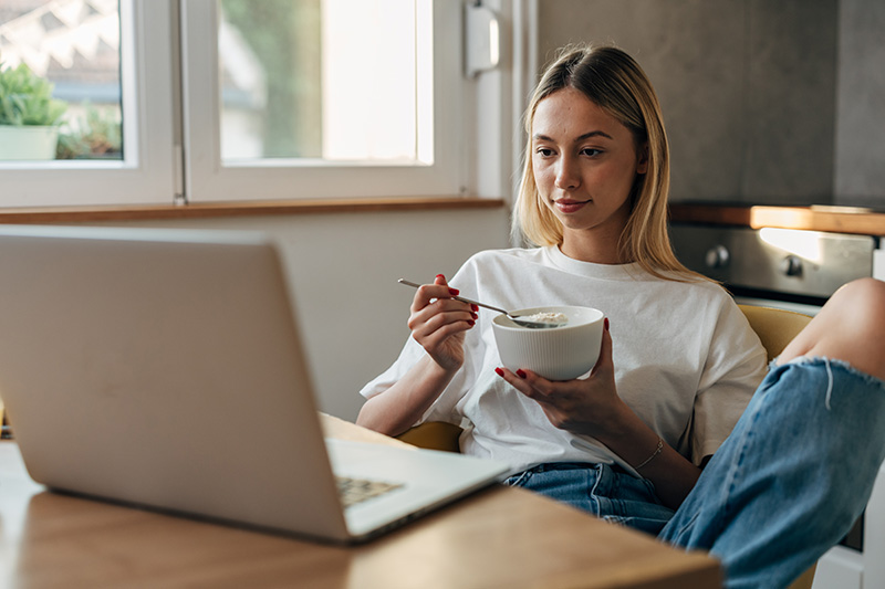 A young blonde woman holds a bowl of oatmeal with a spoon while seated in front of a laptop at a kitchen table