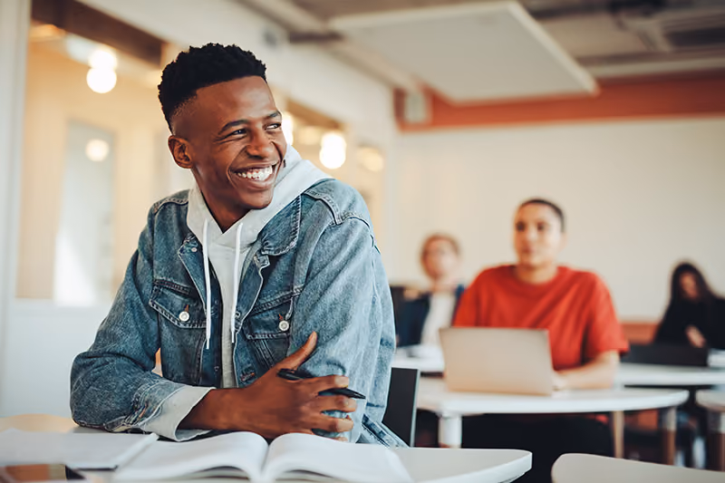 A young man in a jean jacket and hoodie smiles while sitting at a desk in a classroom, looking to his left and holding a pen