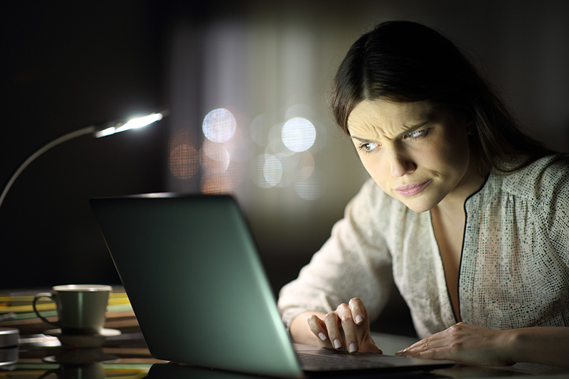 A woman with a suspicious expression looks at a laptop in a dimly lit room.