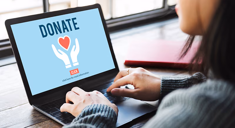 A person types at a computer. The screen is blue with the word "Donate" at the top and a logo of two hands and a heart below