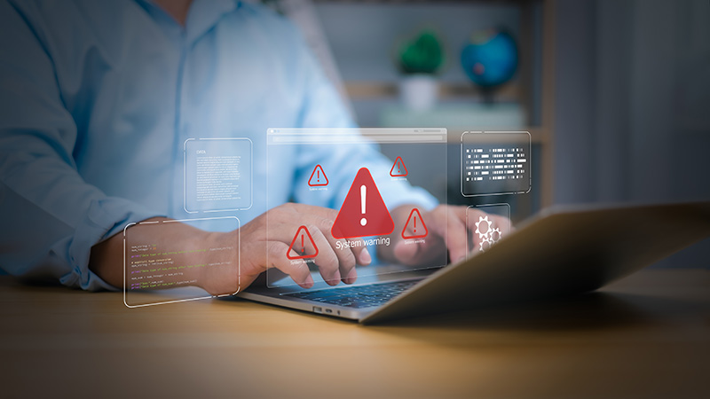 The torso of a person in a blue shirt typing at a laptop with hazard symbols above it