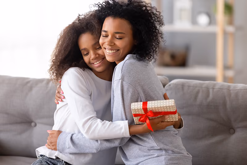 Two woman hug each other while sitting on a couch, the younger one holding a gift