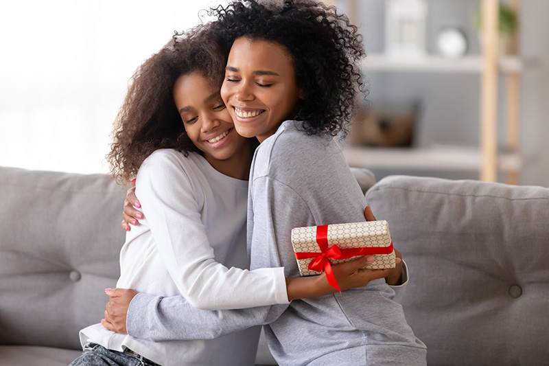 Two woman hug each other while sitting on a couch, the younger one holding a gift