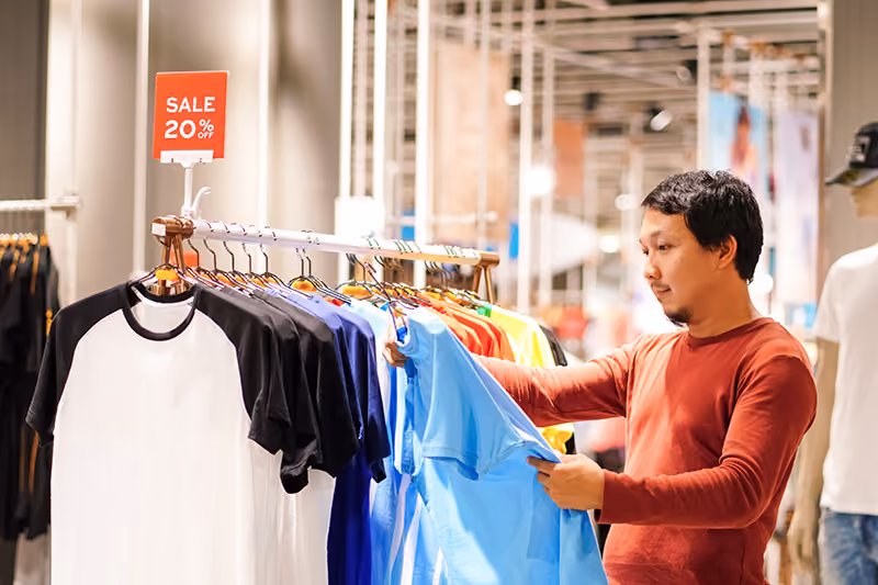 A man with dark hair in a store examining a blue shirt on a rack with a 20% off sale sign