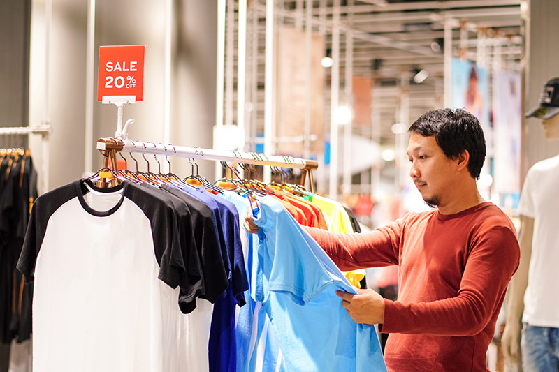 A man with dark hair in a store examining a blue shirt on a rack with a 20% off sale sign