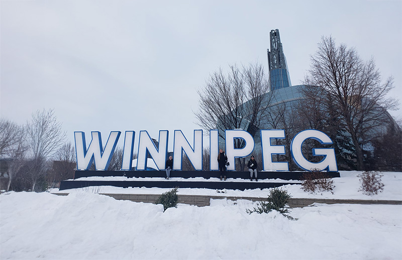 A large "Winnipeg" sign front of a building and trees in winter