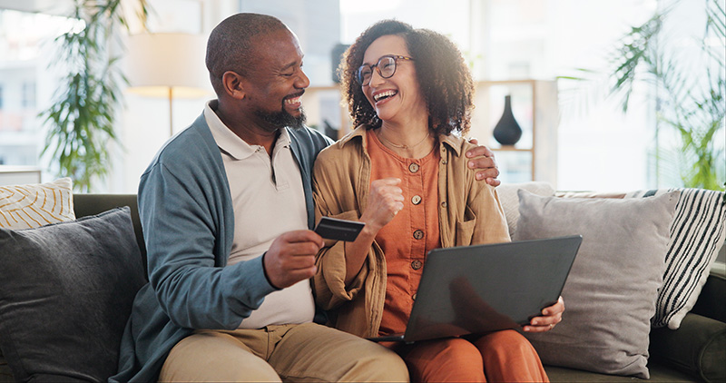 A man, holding a credit card, and a woman sit on a couch with a laptop, laughing and looking at each other