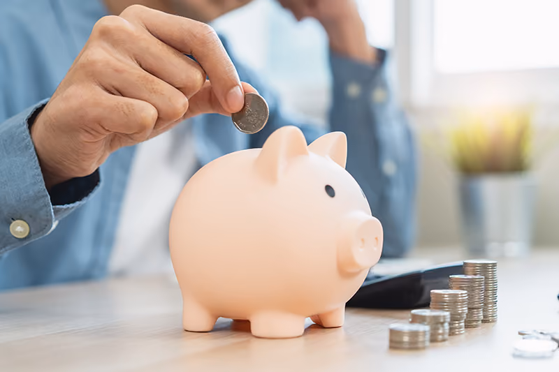 A hand holds a coin, poised to drop it in a pink piggy bank, next to a stack of other coins