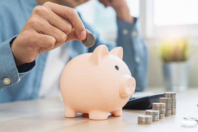 A hand holds a coin, poised to drop it in a pink piggy bank, next to a stack of other coins