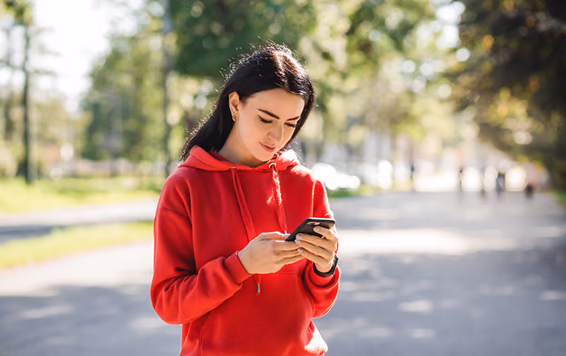 A brunette in a red hoodie looks down at a cellphone, holding it with both hands
