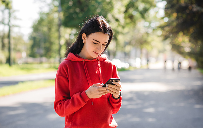 A brunette in a red hoodie looks down at a cellphone, holding it with both hands