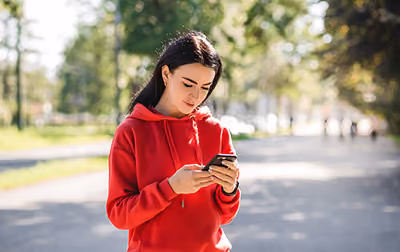 Young woman in a red hoodie looking at her smartphone outdoors on a sunny day.