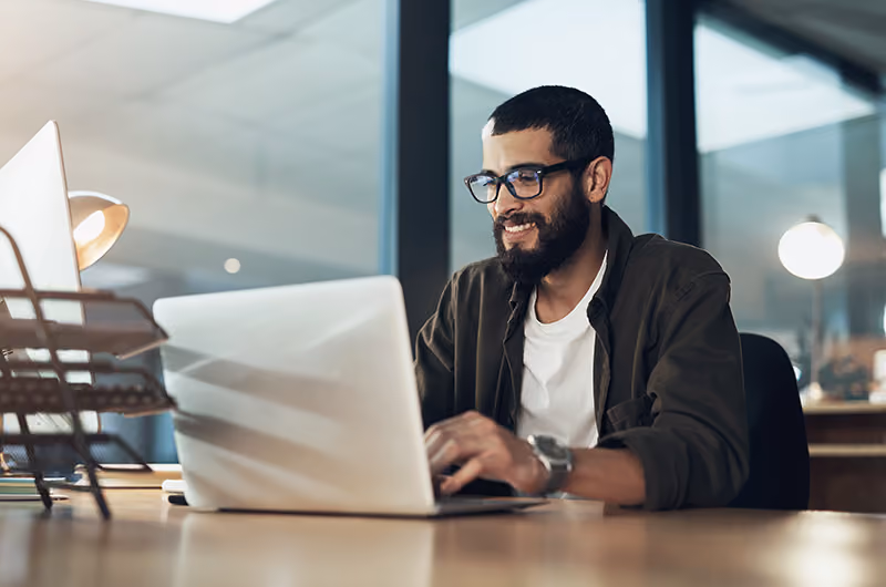 A man with a beard and glasses smiles at a laptop at a table