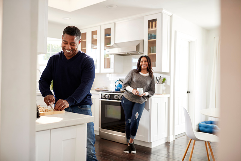 A couple stands in a kitchen, the man using a cutting board and the woman leaning on the counter