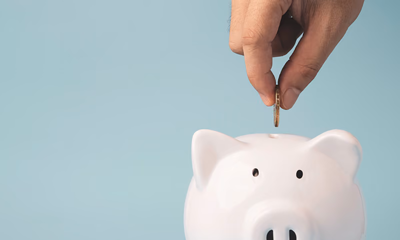 A hand drops a coin in a white piggy bank against a light blue background