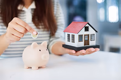 Person putting a coin in a piggy bank, holding a miniature model house in the other hand.