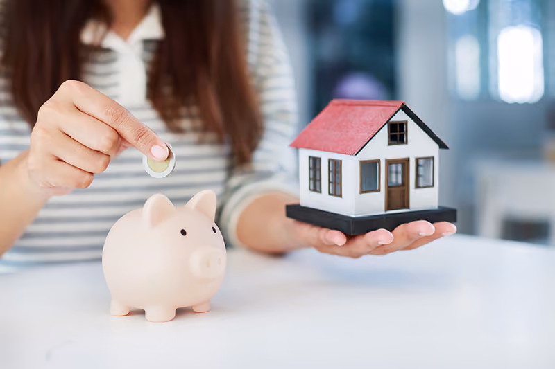 A hand holds a coin above a piggy bank with a mini house model held in the other hand