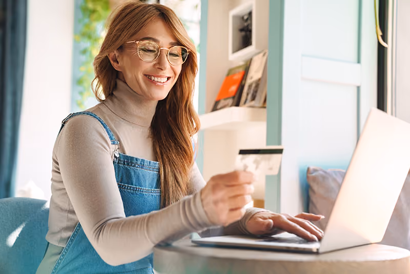 A woman with glasses smiles at a credit card in front of a laptop