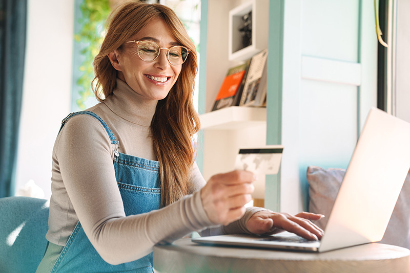A woman with glasses smiles at a credit card in front of a laptop