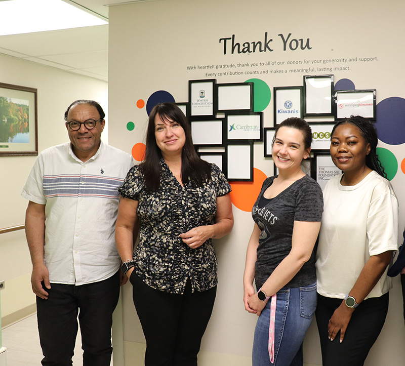 Four people, one man and three women, stand smiling in front of a wall of plaques.